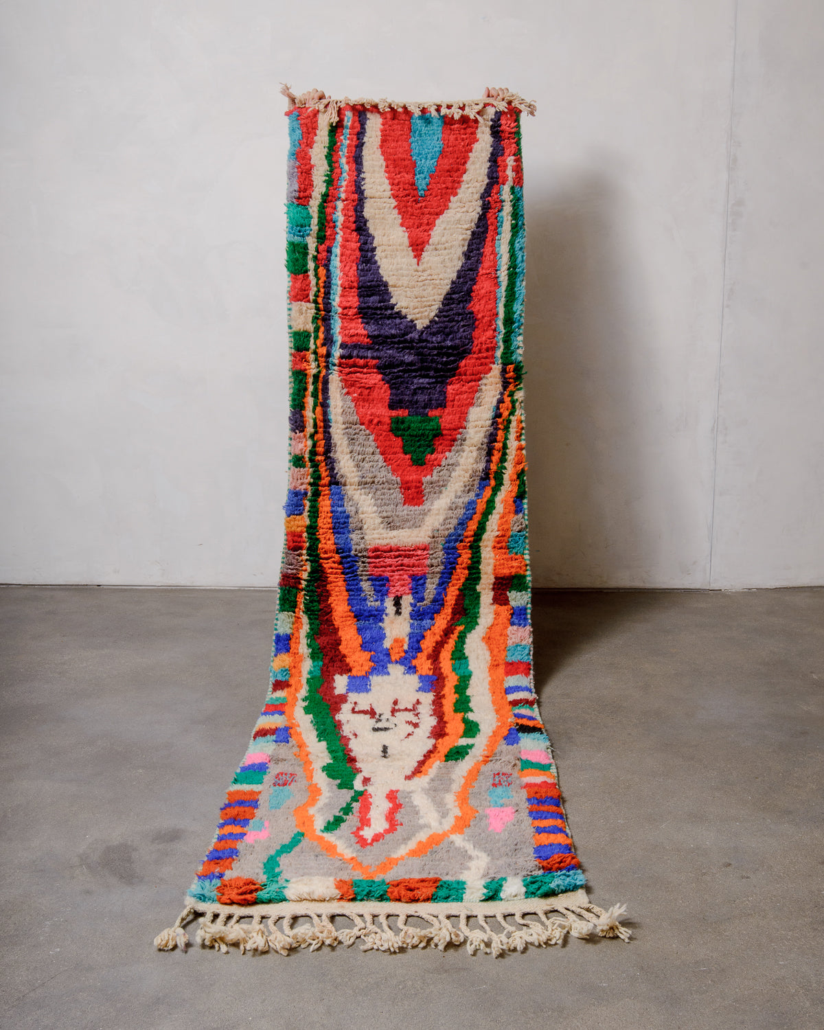 Berberlin vintage runner rug made of brightly coloured wool, being held up in front of a white wall and concrete floor.